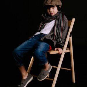 A young boy in trendy clothing poses on a wooden chair against a dark background.