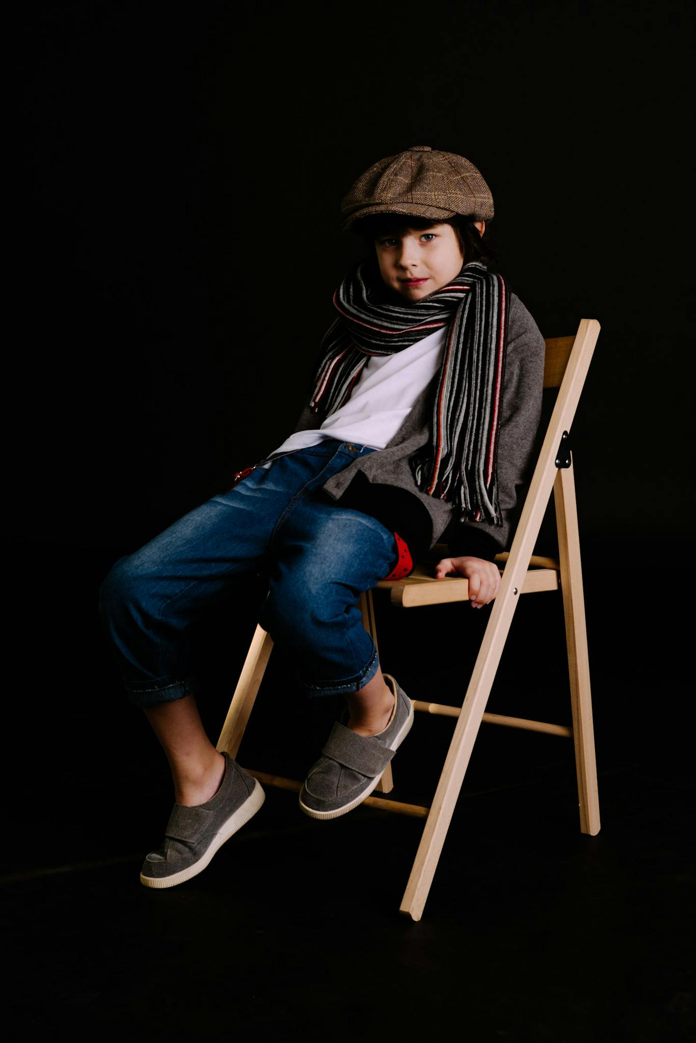 A young boy in trendy clothing poses on a wooden chair against a dark background.