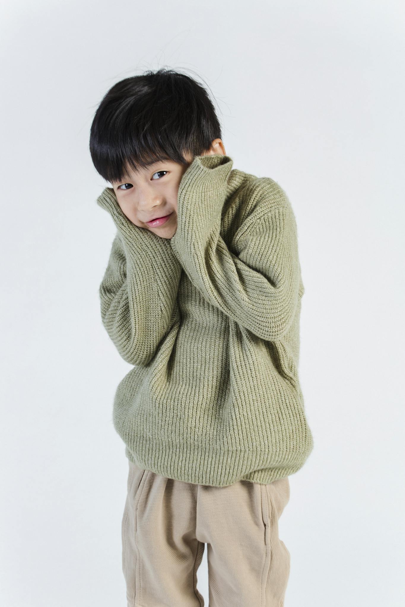 Charming child wearing a cozy green sweater, standing shyly against a white background.
