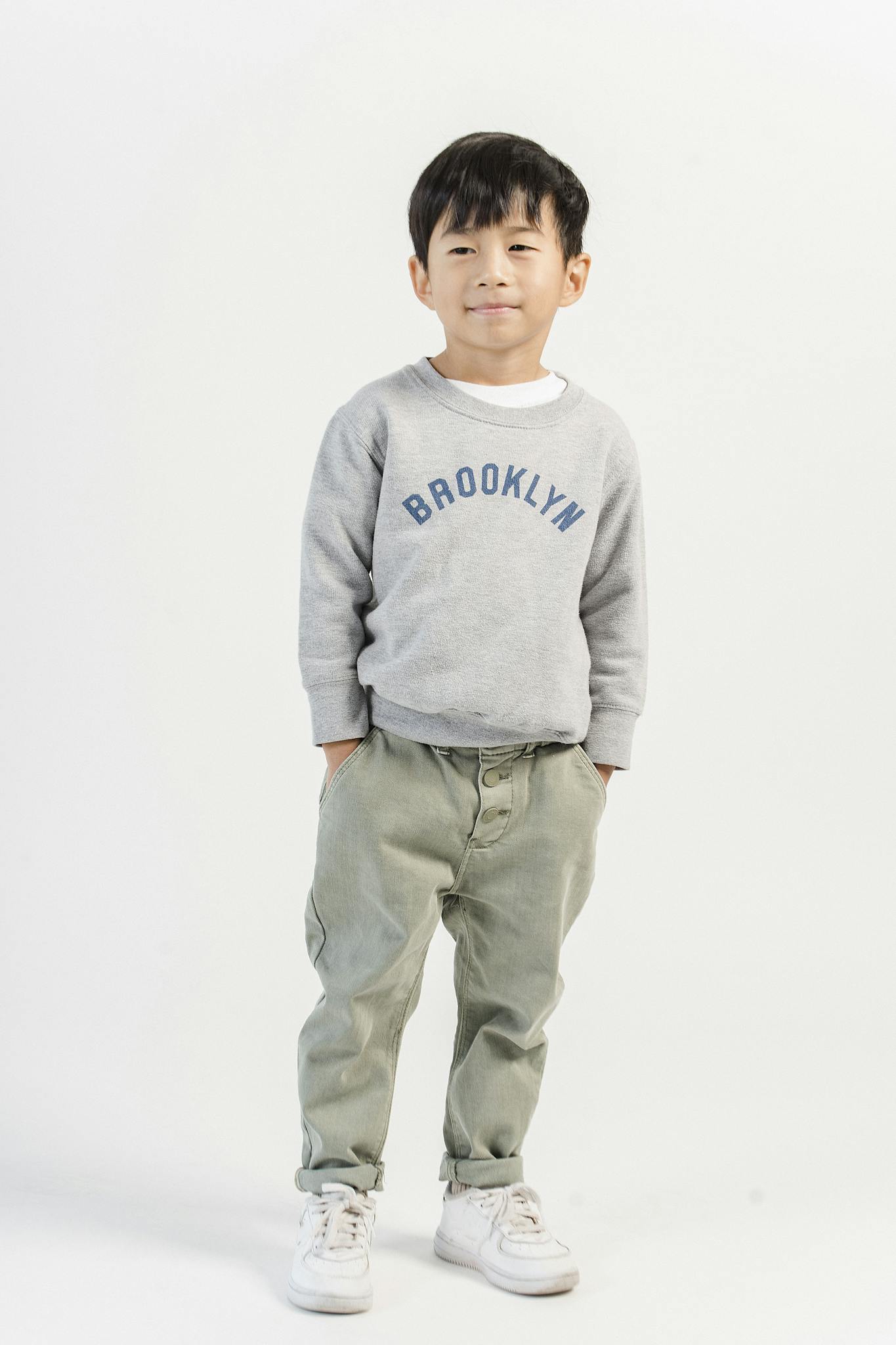 Full body of adorable Asian boy with hands on pockets wearing casual jeans and sweatshirt with sneakers in white studio