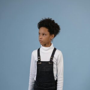 Portrait of a young boy with afro hairstyle wearing denim overalls and turtleneck, studio shot.