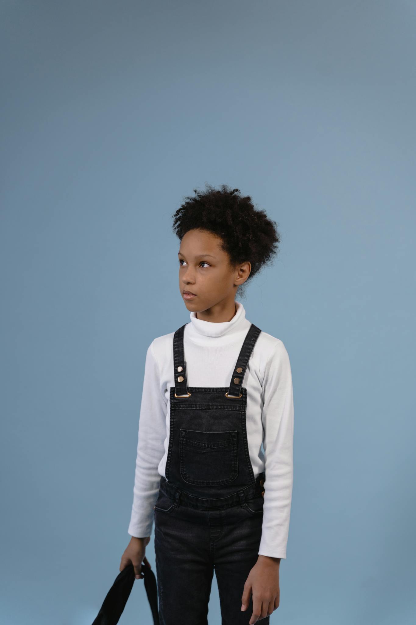 Portrait of a young boy with afro hairstyle wearing denim overalls and turtleneck, studio shot.