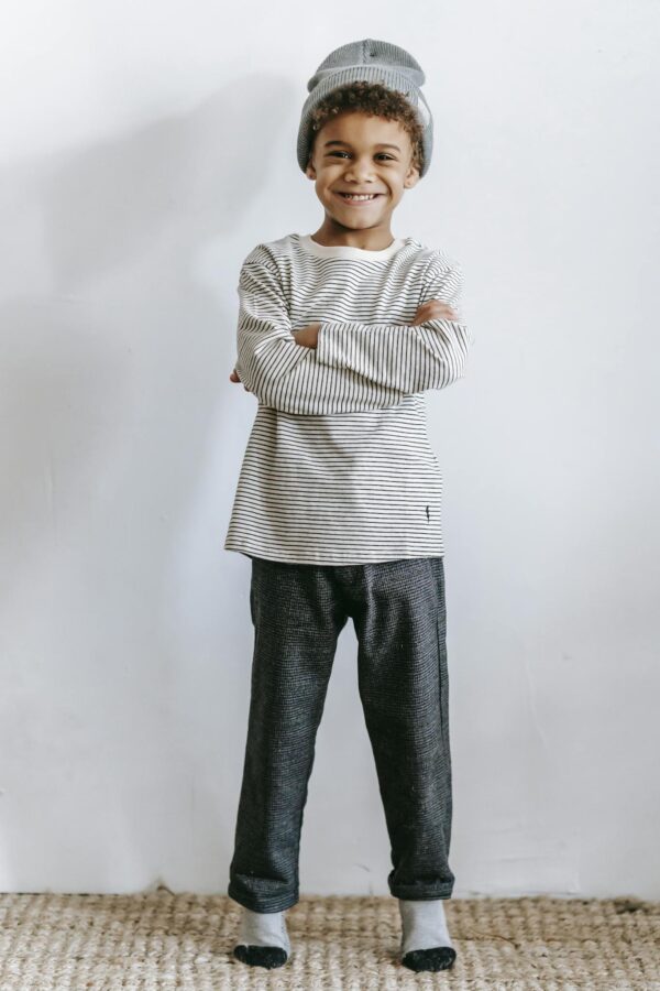 Smiling child in casual striped outfit and beanie, arms crossed, standing indoors.