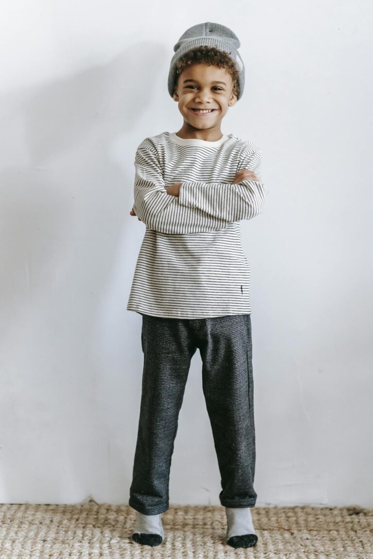 Smiling child in casual striped outfit and beanie, arms crossed, standing indoors.