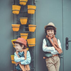 Two boys in fashionable attire pose by a wall with yellow pots, showcasing playful style.