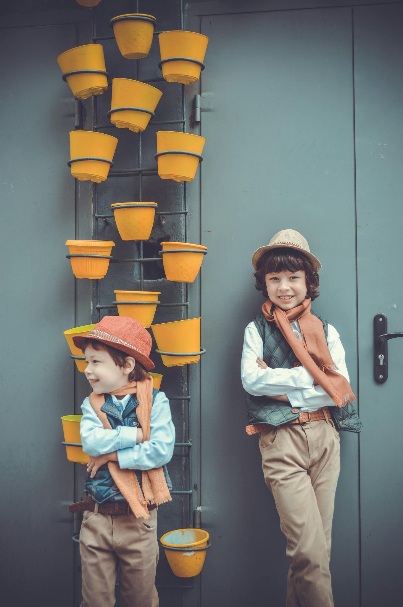 Two boys in fashionable attire pose by a wall with yellow pots, showcasing playful style.
