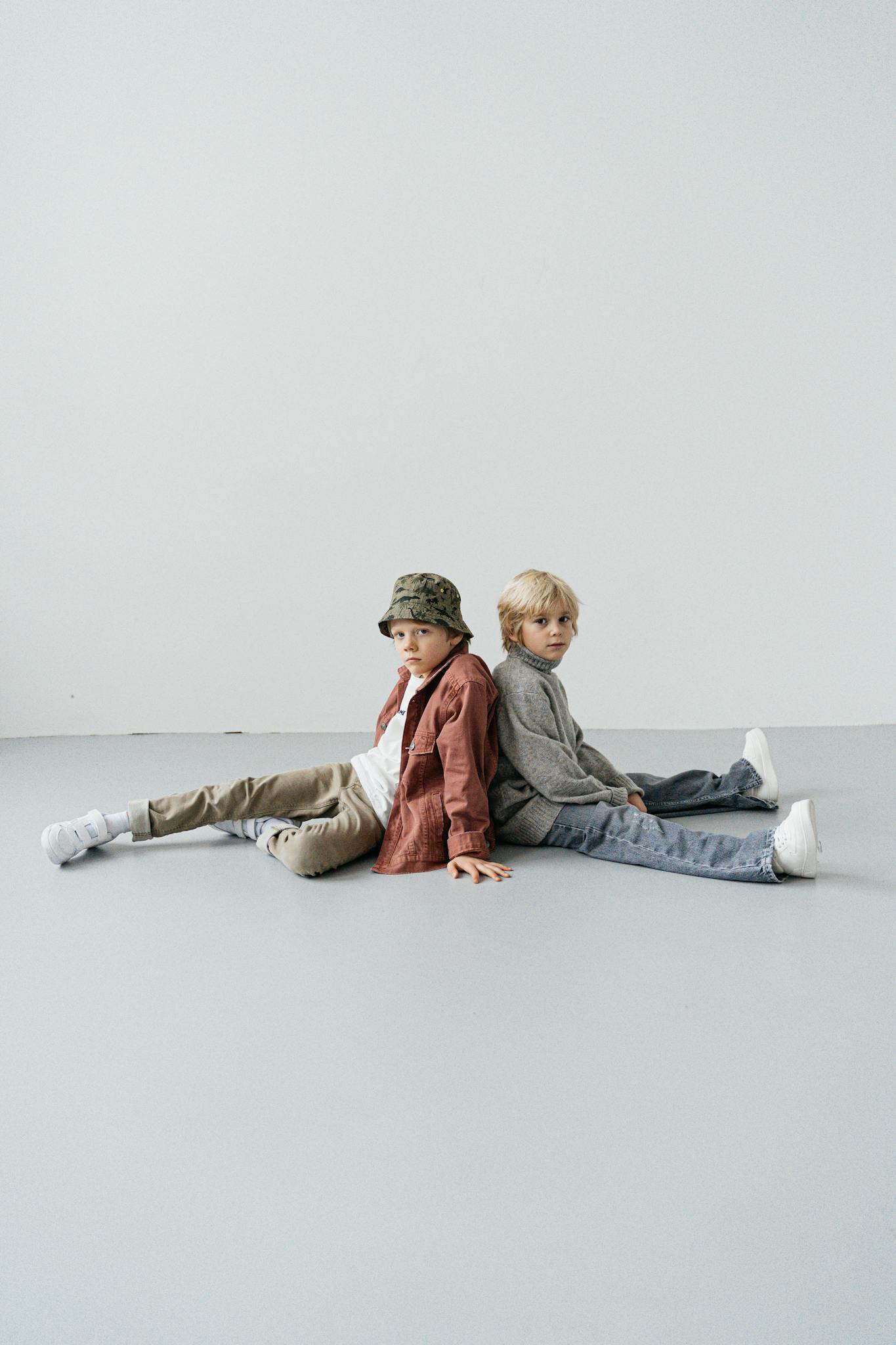 Two young boys in trendy outfits sitting back to back in a minimalistic indoor setting.